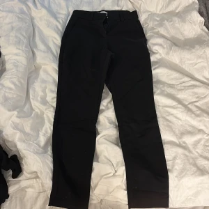 Svarta kostymbyxor i storlek 34 - Svarta kostymbyxor med raka ben och hällor för bälte. Byxorna har sidfickor fram och fuskfickor bak. Materialet är en blandning av polyester, bomull och elastan vilket ger en skön och lite stretchig känsla. Perfekta för en clean och stilren look.