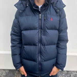 Ralph Lauren jacka  - Snygg mörkblå dunjacka från Polo Ralph Lauren med klassisk röd logga på bröstet. Jackan har hög krage, dragkedja framtill och flera praktiska fickor. Perfekt för kalla dagar och har en stilren, puffig design som ger en modern look.