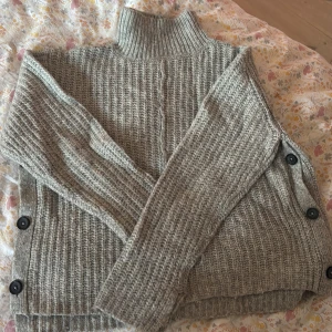 Grå stickad tröja med knappar från Samsa - Säljer en chunky grå stickad tröja från Samsa Collection. Tröjan har hög krage, långa ärmar och coola knappar längs sidorna. Perfekt för lager-på-lager och chill höst- eller vinterstil. Supermjuk och varm, passar dig som gillar en lite loose fit. Det finns även något litet hål på den men det syns knappt