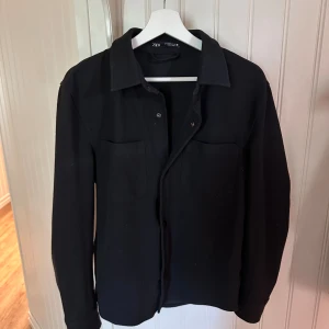 Svart overshirt från Zara - Stilren svart overshirt från Zara med klassisk krage och två bröstfickor. Skjortan har långa ärmar och knäppning framtill. Perfekt att slänga över en t-shirt för en clean look. Materialet känns mjukt och lite tjockare, vilket ger en schysst känsla.