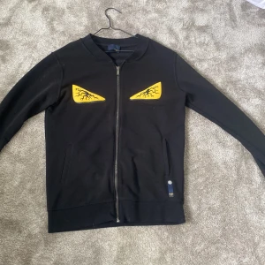 Svart bomberjacka från Fendi med ögon bra skick  - Svart bomberjacka från Fendi med dragkedja framtill och två fickor. På bröstet finns två gula ögon med svarta ådror som ger jackan en unik och edgy vibe. Ribbad krage och muddar. Perfekt för dig som vill sticka ut med en designerjacka.!!! ÄKTA VARA!