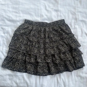 Svart blommig volangkjol By Clara - Supersöt svart kortkjol från By Clara med små vita och beige blommor. Kjolen har flera volanger som ger en flowy och lekfull look. Perfekt för dig som gillar romantiska detaljer och vill ha en kjol som sticker ut lite extra. #blommigt #svartkjol #floralmönster #blackskirt #bohostyle #feminine #kjol #volangkjol #minikjol #sommarkjol #casualstyle