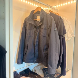 Field jacket från J.Lindeberg - Snygg mörkblå vindjacka från J.Lindeberg med klassisk krage, fyra framfickor med knappar och logotyp på ärmen. Jackan har dragkedja och knappar framtill samt justerbara detaljer i midjan. Perfekt för dig som gillar stilren och sportig look. 