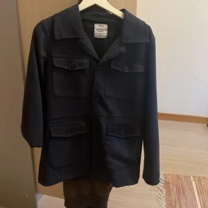 Svart rock från Zoom, slim fit M - Stilren svart rock från Zoom i slim fit-modell. Jackan har fyra stora fickor med lock och knappar framtill samt klassisk krage. Perfekt för dig som gillar en clean och tidlös look. Passar dig som vill ha en snygg och enkel jacka till vardags.