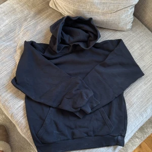 Svart hoodie från H&M med huva - En svart hoodie från H&M i mjuk bomull med klassisk huva och känguruficka framtill. Tröjan har långa ärmar med ribbade muddar och en relaxed passform. Perfekt för chill dagar eller när du vill ha en enkel och stilren look.