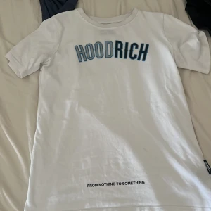 Vit HOODRICH t-shirt med tryck - Snygg vit t-shirt från HOODRICH med blå och svart text på bröstet och ett mindre tryck längst ner framtill. Klassisk passform med rund hals och korta ärmar. Perfekt för dig som gillar streetwear och vill sticka ut lite extra.