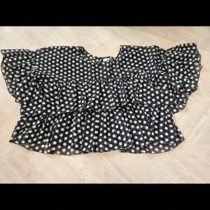Prickig volangblus i svart och vitt - Säljer en svart blus med vita prickar och stora volanger. Blusen har korta, breda ärmar och är tillverkad i ett lätt, transparent material som ger en luftig känsla. Perfekt för dig som gillar statement-plagg med mycket rörelse och cool retrostil.