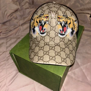 Gucci keps med tigertryck och GG-mönster (FEJK) - Säljer en snygg Gucci keps med klassiskt GG-mönster i beige och brunt, prydd med två coola tigerhuvuden framtill. Kepsen har svart mesh på baksidan för extra ventilation och justerbar passform. Perfekt statement piece för dig som vill sticka ut. 