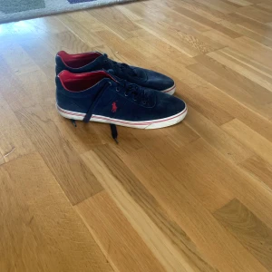 Blå mockasneakers från Polo Ralph Lauren - Snygga sneakers i mörkblå mocka från Polo Ralph Lauren med röd insida och röd logga på sidan. Skorna har vita sulor med röda detaljer och klassisk snörning. Perfekta för dig som gillar stilrena och sportiga skor med en touch av färg.