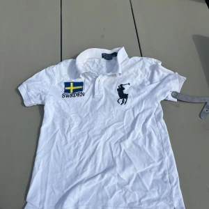 Vit Polo t-shirt från Ralph Lauren - Vit kortärmad t-shirt från Polo Ralph Lauren med klassisk pikékrage och knappar. På bröstet finns en stor broderad Polo-logga i marinblått och en svensk flagga med texten SWEDEN. Materialet är mjuk bomull och passformen är custom fit.