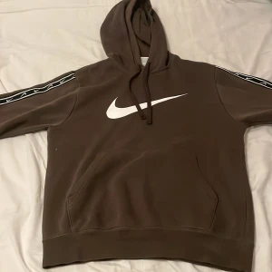  Nike hoodie, om ni vill ha med byxorna kan ni köpa separat i min andra annons - Säljer en brun hoodie från Nike med stor vit swoosh-logga på bröstet och svarta band med små vita swooshes längs ärmarna. Tröjan har huva med snörning, känguruficka och är långärmad. Perfekt för dig som gillar sportig och chill stil.