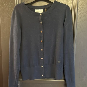 Mörkblå kofta från Holly & White - En klassisk mörkblå kofta från Holly & White med rund halsringning och knappar framtill. Långärmad och i mjukt material som passar perfekt till lager-på-lager. Enkel och stilren design med diskret logga vid nederkanten.