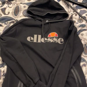 Svart hoodie från Ellesse med logga - Svart hoodie från Ellesse i storlek M med stor vit och orange logga på bröstet. Tröjan har huva med snörning och långa ärmar. Perfekt för en chill och sportig stil. Materialet känns mjukt och bekvämt.