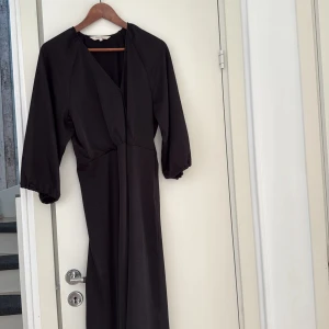 Svart omlottklänning från Part Two - Snygg svart långklänning från Part Two med omlott-design och v-ringad hals. Klänningen har trekvartsärmar med lätt puff och resår vid ärmslut. Materialet är mjukt och har ett elegant fall, perfekt för dig som gillar stilrena och tidlösa plagg.