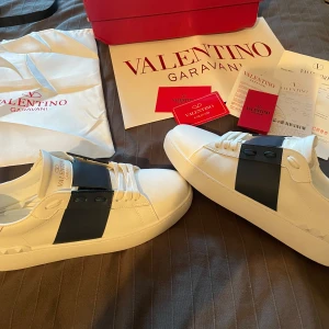 Valentino Garavani Open sneakers vit/svart - Snygga Valentino Garavani Open sneakers i vitt skinn med bred svart rem över vristen. Klassisk rund tå, vita snören och coola nitar på hälen. Perfekta för dig som vill ha en stilren men ändå lyxig look. Stolek 42! 