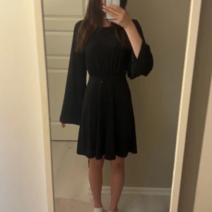 Svart kortklänning med öppen rygg - Snygg svart kortklänning med vida långärmar och markerad midja. Klänningen har en öppen rygg med knytband upptill, den är knappt använd.