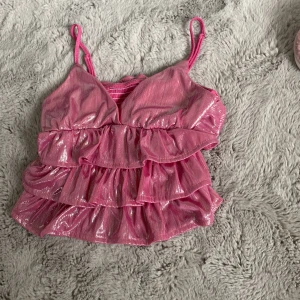 Glittrig rosa volang-linnetopp - Supersöt rosa linnetopp med glittrigt tyg och flera volanglager. Smala axelband och v-ringad framtill, perfekt för dig som vill sticka ut. Toppen har en lätt skimrande finish och elastisk rygg för extra komfort.