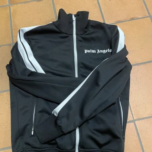 Palm Angels svart track jacket - Svart track jacket från Palm Angels med vita ränder längs ärmarna och vit dragkedja. Jackan har hög krage, två fickor med dragkedja och logga på bröstet. Perfekt för en sportig och street-inspirerad look. Den är äkta köpt länge sen ej kvitto kvar 