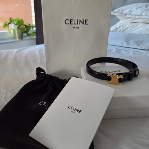 Svart Celine Triomphe bälte i skinn 18mm - Snyggt svart bälte från Celine med ikoniskt Triomphe-spänne i guld. Bältet är gjort i äkta skinn och har en stilren, smal design som ger en lyxig känsla. Perfekt accessoar för att lyfta vilken outfit som helst med exklusiv vibe. Nypris mellan 5200-5700kr.