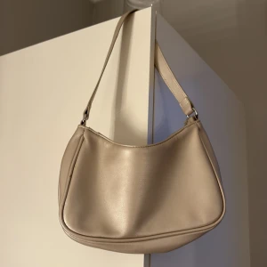 Beige axelväska i skinnimitation - Snygg beige axelväska med minimalistisk design och mjuka former. Väskan har ett stilrent utseende och är tillverkad i skinnimitation med en bred axelrem. Perfekt för att bära dina viktigaste grejer och passar till många olika outfits.