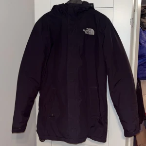 Svart parkajacka från The North Face - Säljer en svart parkajacka från The North Face i storlek M. Jackan har huva, dragkedja och två fickor framtill. Perfekt för kyliga dagar och har en stilren look med logga på bröstet och ryggen. Materialet är slitstarkt och jackan är långärmad.