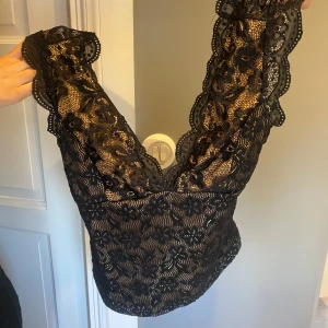 Svart spetsig croppad blus med öppen rygg - Supersnygg svart blus i spets med blommigt mönster och djup v-ringning. Toppen är croppad och har öppen rygg, vilket ger en riktigt trendig vibe. Perfekt för dig som vill sticka ut med coola detaljer och romantisk känsla.