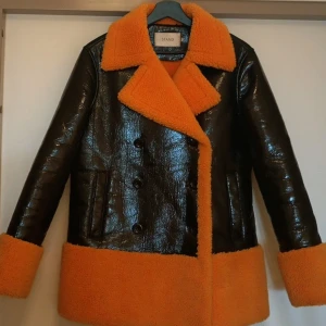 Svart och orange pälsjacka från Stand - Unik pälsjacka från Stand Studioi svart och orange. Yttermaterialet är glansigt svart med orange teddyfoder, krage och muddar. Dubbelknäppt framtill med stora knappar och två fickor. Jackan har en loose passform och är perfekt för dig som vill sticka ut.Jackn är i nyskick och förvarad i kostymväska. Size 36 men jag har 38/40 och den sitter snyggt ,när man bär den så är ............... All eyes on you!