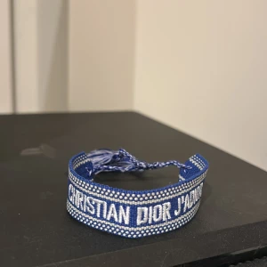 Blått armband från Christian Dior - Snyggt blått tygarmband från Christian Dior med texten 'CHRISTIAN DIOR J'ADIOR' broderad i vitt. Armbandet har justerbar knytning med två tofsar i blått och vitt. Perfekt accessoar för att lyfta din outfit med en lyxig touch.
