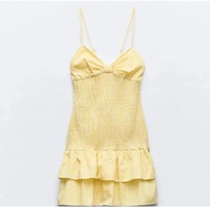 Gul kort klänning med volang - Söt gul kort klänning med tunna axelband och volang nertill. Klänningen har ett diskret randigt mönster och en feminin passform med lätt rynkning vid bysten. Perfekt för soliga dagar och sommarhäng med vänner.