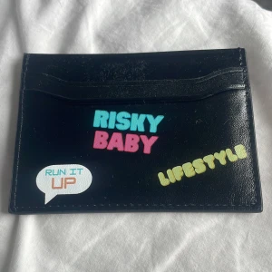 Svart korthållare från Risky - Svart korthållare i skinn med färgglada tryck som 'RISKY BABY', 'LIFESTYLE' och 'RUN IT UP'. Plats för flera kort på båda sidor och präglad logga på baksidan. Perfekt för dig som vill ha en unik och personlig accessoar i fickan. Helt ny. Nypris 386kr 