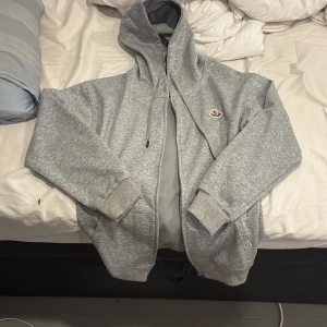 Grå Moncler hoodie med dragkedja - Snygg grå hoodie från Moncler med dragkedja framtill, huva med snörning och klassisk logga på bröstet. Tillverkad i mjuk bomullsmix, perfekt för chill dagar. Ribbstickade muddar och två fickor framtill ger en clean och sportig look.