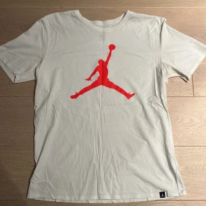 Vit Jordan t-shirt med röd logga - Snygg vit t-shirt från Jordan med den klassiska röda Jumpman-loggan tryckt stort på bröstet. T-shirten har rund hals, korta ärmar och är gjord i mjuk bomull. Perfekt för dig som gillar sportig och clean stil.
