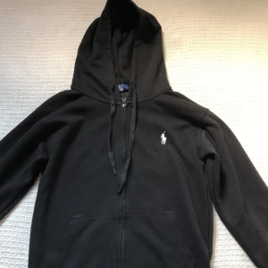 Svart hoodie från Polo Ralph Lauren - Svart hoodie från Polo Ralph Lauren med klassisk vit broderad logga på bröstet. Tröjan har dragkedja, huva med snörning och känguruficka framtill. Tillverkad i mjuk bomull, perfekt för chill dagar eller när du vill ha en clean streetstyle. Använd en gång pågrund av storleken då jag vanligtvis inte använder storlek ”s”