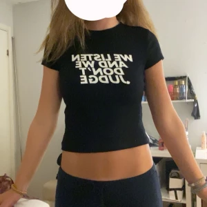 Svart croppad topp med texttryck - Svart croppad topp från Bershka med vit text 'WE LISTEN AND WE DON'T JUDGE' framtill. T-shirten har korta ärmar, rund halsringning och är figurnära i modellen. Perfekt för dig som gillar statement-plagg och enkel stil. 