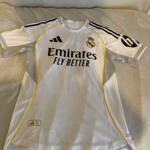 Real Madrid Ronaldo 7 matchtröja Adidas - Säljer en officiell Real Madrid fotbollströja från Adidas med Ronaldo 7-tryck på ryggen. Tröjan är vit med svarta detaljer, gula linjer och klubbmärke på bröstet. Tillverkad i lätt och ventilerande material, perfekt för fotbollsplanen eller som supporterplagg.