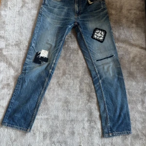 Nudie Jeans blå patchade jeans 29/30 - Unika blå jeans från Nudie Jeans med flera coola patchade detaljer och slitningar på benen. Klassisk femficksmodell med orange sömmar och broderade bakfickor. Jeansen har rak passform och är tillverkade i bomullsdenim. 1 of 1 i världen. Pris går såklart att diskutera!