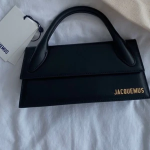 Svart handväska från Jacquemus - Säljer en svart handväska från Jacquemus med stilren, rektangulär form och ett elegant handtag. Väskan är i skinn och har guldigt Jacquemus-logo på framsidan. Perfekt för dig som gillar minimalistisk och trendig design. Design namn Jacquemus Le chiquito long ord.pris 7,638kr