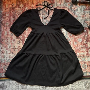 Svart klänning med v-ringning och knyt - Säljer en svart klänning med djup v-ringning och knyt i nacken. Klänningen har puffiga trekvartsärmar och volang i kjolen för en extra flowy känsla. Perfekt för dig som gillar en stilren men ändå trendig look.