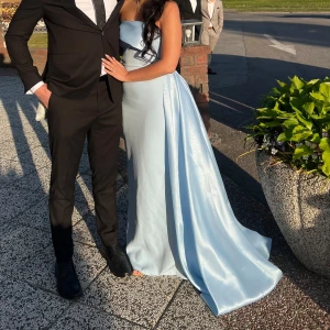 Ljusblå axelbandslös balklänning - Säljer en elegant ljusblå långklänning med axelbandslös design och draperad överdel. Klänningen har en figurnära passform och ett långt släp som ger en extra lyxig känsla. Perfekt för bal eller speciella tillfällen. Använd en gång.