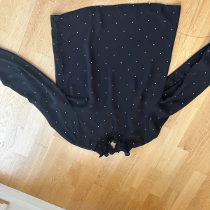 Svart prickig blus  - Supersnygg svart blus med vita prickar och långärmade ärmar. Smockdetaljer vid halsringning och ärmslut ger en cool och trendig look. Blusen har en rak form och är gjord i ett lätt, flowy material som känns skönt mot huden.