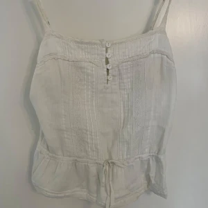 Vit linnetopp med spets och knappar - Söt vit linnetopp med smala axelband, dekorativa spetsdetaljer och små knappar framtill. Toppen har tunna vertikala ränder, volangkant nertill och knytband i midjan. Perfekt för varma dagar och ger en romantisk vibe.