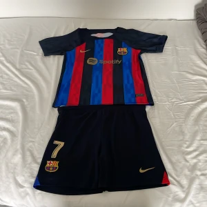 FC Barcelona Dembele matchtröja Nike - Säljer en FC Barcelona matchtröja med shorts, officiell Nike-version med O. Dembélé och nummer 7 på ryggen. Tröjan har klassiska blå och röda vertikala ränder, guldiga detaljer och klubbmärke. Materialet är lätt polyester, perfekt för fotboll. Spotify-logga på bröstet. Storlek 28, passar dom som är mellan ca 155-170 cm