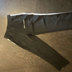 Palm Angels svarta mjukisbyxor med vit stripe - Svarta mjukisbyxor från Palm Angels med vit sidoremsa och broderad logga på låret. Byxorna har elastisk midja och raka ben, perfekta för en chill och sportig look. Materialet är mjukt och följsamt, vilket gör dem riktigt sköna att bära.