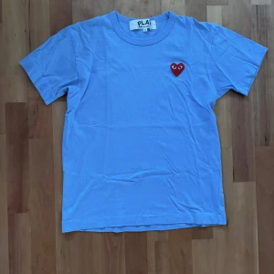 Blå Comme des Garçons PLAY t-shirt - Blå t-shirt från Comme des Garçons PLAY i bomull med klassisk passform. På bröstet sitter det ikoniska röda hjärta med ögon. T-shirten har rund hals och korta ärmar, perfekt för en clean och chill look.