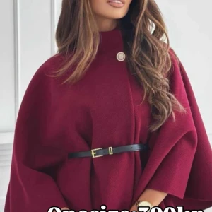 Vinröd kappa med bälte och hög krage - Stilren vinröd kappa med hög krage och elegant svart bälte i midjan. Kappan har en snygg cape-liknande form och stängs med en dekorativ knapp upptill. Perfekt för dig som vill sticka ut med en klassisk men modern look.