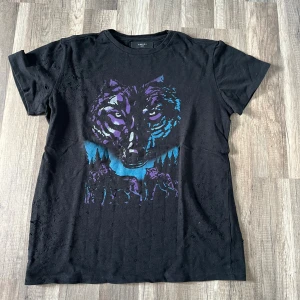 Svart t-shirt med vargmotiv från Amiri - Svart t-shirt från Amiri med ett stort tryck av en varg i lila och blå toner på bröstet. T-shirten har korta ärmar och rund hals. Materialet är mjuk bomull och passformen är normal. Perfekt för dig som gillar coola djurmotiv och streetwear.