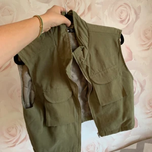Olivgrön quiltad väst från SHEIN - Snygg olivgrön väst från SHEIN i storlek M. Croppad modell med två stora fickor framtill och dragkedja. Quiltad insida i beige och hög krage som ger en cool look. Perfekt att slänga över en t-shirt för extra stil.