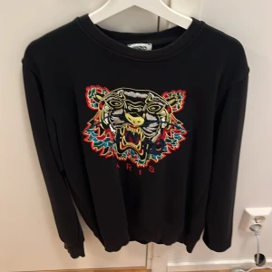 Svart Kenzo sweatshirt med tigerbrodyr - Cool svart sweatshirt från Kenzo med ikonisk färgglad tigerbrodyr och texten 'Paris' framtill. Tröjan har rund halsringning och långa ärmar. Perfekt statement-plagg för dig som vill sticka ut med stil.