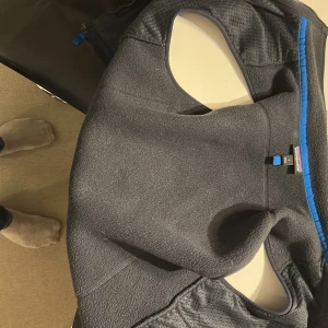 Mörkgrå fleeceväst från Patagonia - Säljer en mörkgrå fleeceväst från Patagonia i storlek S. Västen har blå detaljer vid dragkedjorna och hög krage. Perfekt att slänga över en hoodie eller långärmad tröja. Klassisk Patagonia-logga på bröstet och flera praktiska fickor med dragkedja.