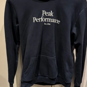 Mörkblå hoodie från Peak Performance - Snygg mörkblå hoodie från Peak Performance med vit broderad logga på bröstet. Klassisk känguruficka framtill, huva och långa ärmar. Bra begagnat skick!
